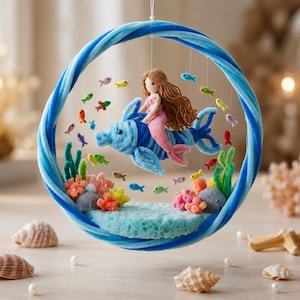 Peut inclure: Un mobile suspendu circulaire avec un cadre en cerceau enveloppé de velours bleu. À l'intérieur, une sirène en feutrine aux cheveux bruns chevauche un dauphin en feutrine bleue. Le décor inclut des petits poissons, des algues et du corail.