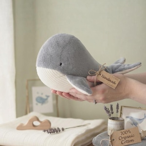 Peut inclure: Une peluche baleine douce de couleur grise et crème avec un corps arrondi et un petit œil noir. Une étiquette en papier brun attachée avec de la ficelle à la nageoire de la baleine porte le mot Handmade. La peluche a un dessous côtelé.