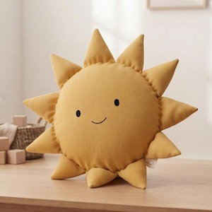 Peut inclure: Un coussin jaune moutarde en forme de soleil avec un visage brodé simple composé de deux petits yeux noirs circulaires et d'une fine ligne courbe pour la bouche. Le coussin présente douze rayons pointus autour du centre circulaire.