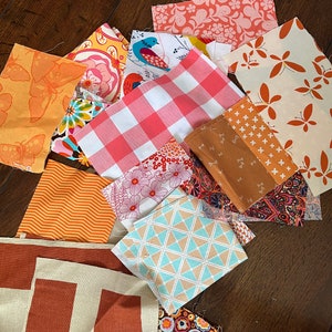 May include: A collection of fabric squares in various patterns and colors, including orange, pink, white, and brown. The fabrics feature geometric designs, floral patterns, and abstract prints.