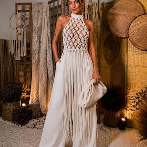 Op de afbeelding: Een crèmekleurige jumpsuit met een macramé haltertop met een ruitpatroon, een hoge halslijn en lange franjes bij de taille. Het kledingstuk bevat een broek met wijde pijpen, zijzakken en een bijpassende crèmekleurige clutch.