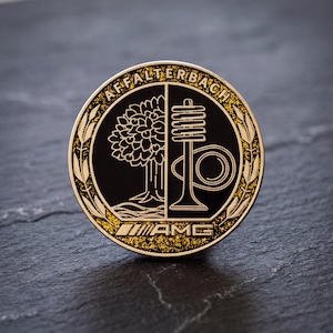 Könnte beinhalten: Ein runder Metall-Pin mit schwarzer Mitte, die eine goldene Baum- und Ventilgrafik zeigt. Der Schriftzug AFFALTERBACH ist oben bogenförmig angebracht, das AMG-Logo befindet sich unten. Ein goldener Glitzerrand umgibt die Mitte, eingerahmt von einem Ährenmuster.
