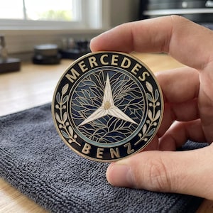 Può includere: Un emblema metallico circolare con il logo Mercedes-Benz, caratterizzato da una stella a tre punte bianca su uno sfondo blu scuro con intricati motivi a foglia. Il bordo dorato riporta la scritta MERCEDES BENZ in nero.