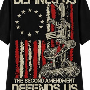 May include: A black t-shirt featuring a graphic of a coiled rattlesnake and a rifle superimposed over a distressed American flag. The text reads THE FIRST AMENDMENT DEFINES US at the top and THE SECOND AMENDMENT DEFENDS US at the bottom.
