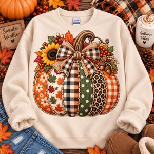 May include: A cream-coloured crewneck sweatshirt featuring a large graphic of a pumpkin decorated with various patterns including plaid, polka dots, leopard print, and autumn leaves. A large plaid bow sits atop the pumpkin, surrounded by sunflowers.