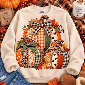 May include: A cream-coloured crewneck sweatshirt featuring a graphic of three pumpkins with patchwork patterns including gingham, leopard print, hearts, and flowers. Each pumpkin is adorned with a decorative bow, autumn leaves, and berries.