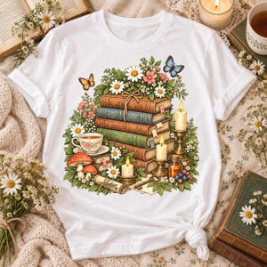 May include: A white short-sleeve t-shirt featuring a graphic print of a stack of vintage books topped with daisies and butterflies. The illustration includes a teacup, lit candles in holders, red mushrooms, and a small glass bottle.