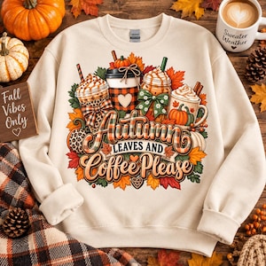 May include: A cream-coloured crewneck sweatshirt featuring a graphic print of three coffee cups surrounded by autumn leaves and pumpkins. The text reads Autumn Leaves and Coffee Please. The design includes plaid patterns, leopard print, and bows.