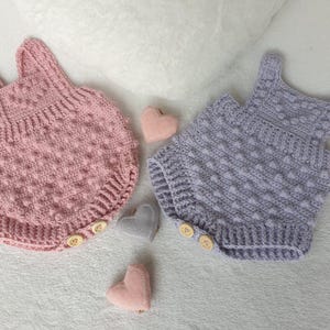 Puede incluir: Dos peleles de bebé tejidos a ganchillo, uno en rosa empolvado y otro en lavanda claro, con un patrón de puntos de burbuja texturizados, tirantes anchos y dos pequeños botones de madera en las aberturas de las piernas, junto a varios corazones de fieltro.
