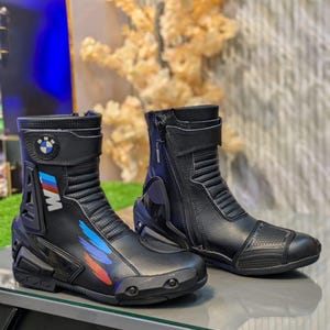 Puede incluir: Un par de botas de moto negras con el logotipo de BMW en la correa del tobillo, cierre de cremallera lateral y detalles gráficos en azul, morado y rojo en el lateral. Las botas incluyen secciones reforzadas en el talón y la puntera.