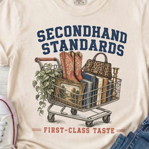 Può includere: Una t-shirt color crema con la grafica di un carrello della spesa in metallo pieno di una pianta in vaso, un paio di stivali da cowboy marroni, una borsa leopardata, diversi libri e un quadro paesaggistico, con il testo SECONDHAND STANDARDS FIRST-CLASS TASTE.