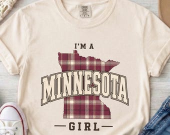 May include: A cream-colored short-sleeve t-shirt featuring a maroon and white plaid graphic of the state of Minnesota. The text I'M A MINNESOTA GIRL is printed in dark brown block lettering across the center of the state outline.