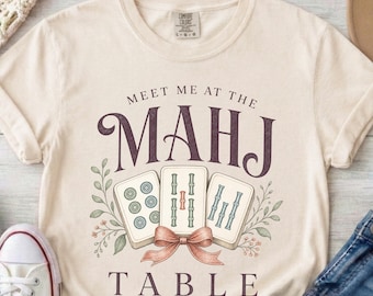 以下が含まれることがあります： 「MEET ME AT THE MAHJ TABLE」という文字がプリントされたクリーム色の半袖Tシャツ。デザインには3枚の麻雀牌、装飾的なリボン、そして落ち着いた緑、赤、青の色調で描かれた植物のイラストがあしらわれています。