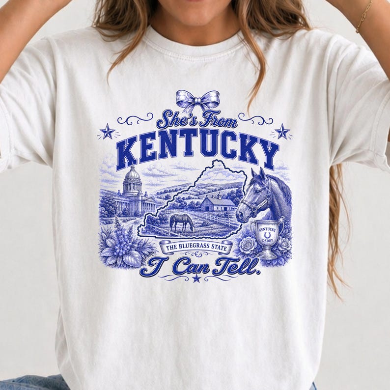 A white t-shirt with a graphic design featuring a horse, flowers, and a state map, along with the text "She's From Kentucky" and "I Can Tell."