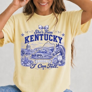 A woman wearing a yellow t-shirt with a graphic design featuring a horse, flowers, and text related to Kentucky.