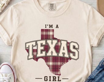 以下が含まれることがあります： クリーム色の半袖Tシャツ。テキサス州の形をしたマルーンと白のチェック柄グラフィックが特徴です。州の形の上に「I'M A」という文字があり、その上に大きなブロック体で「TEXAS」とプリントされ、その下に「GIRL」という文字が配置されています。