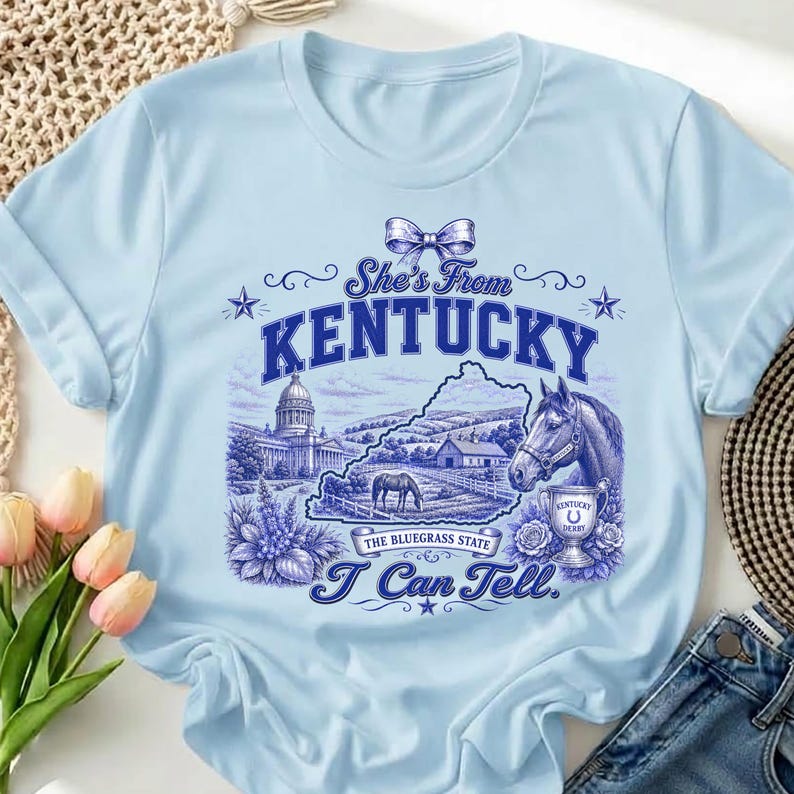 A light blue t-shirt with a graphic design depicting a horse, buildings, and flowers, along with the text "She's From Kentucky" and "I Can Tell."