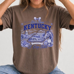A woman wearing a brown t-shirt with a graphic design featuring a horse, buildings, and text related to Kentucky.