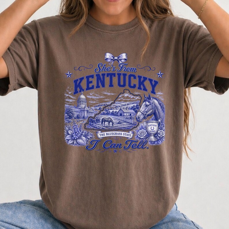 A woman wearing a brown t-shirt with a graphic design featuring a horse, buildings, and text related to Kentucky.