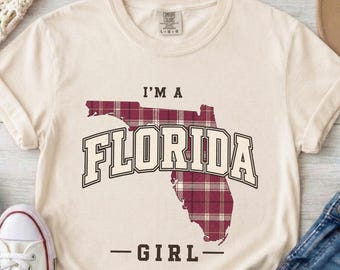 以下が含まれることがあります： クリーム色の半袖Tシャツで、フロリダ州の形をしたマルーンと白のチェック柄グラフィックが特徴です。地図の上に「I'M A」という文字があり、その下に「FLORIDA GIRL」が太字のダークブラウンのカレッジ風フォントでプリントされています。