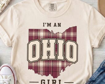 May include: A cream-colored short-sleeve t-shirt featuring a maroon and white plaid map of Ohio. The text I'M AN OHIO GIRL is printed in dark brown block lettering above and below the state shape. The shirt has a crew neckline and a tag.