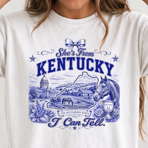 A white t-shirt with a graphic design featuring a horse, buildings, and text related to Kentucky.