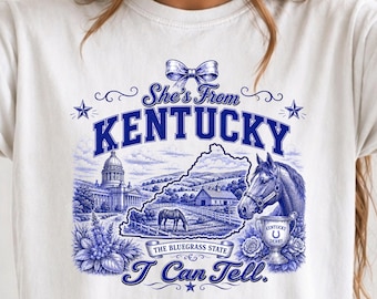 以下が含まれることがあります： 「She's From KENTUCKY I Can Tell.」という文字が青色でプリントされた白いTシャツ。デザインにはケンタッキー州の地図、馬、トロフィー、ドーム型の建物、リボン、そして「THE BLUEGRASS STATE」というフレーズが含まれています。