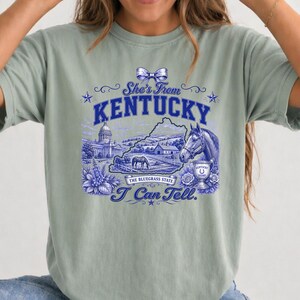 A woman wearing a green t-shirt with a graphic design featuring a horse, flowers, and a map of Kentucky.