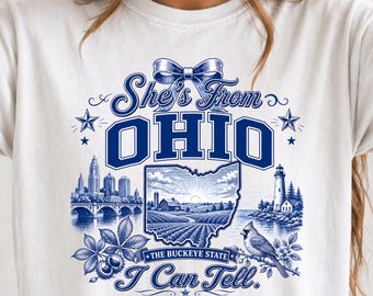 以下が含まれることがあります： 「She's From OHIO I Can Tell」という文字が青色でプリントされた白いTシャツ。デザインにはオハイオ州の地図、都市のスカイライン、灯台、ショウジョウコウカンチョウ、トチノキの葉、リボンが描かれており、「THE BUCKEYE STATE」という文字も含まれています。