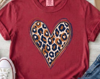 以下が含まれることがあります： 胸元に大きなハートのグラフィックが描かれたダークレッドの半袖Tシャツ。ハートの中は茶色と黒のヒョウ柄で、青、オレンジ、ダークレッドの細いスケッチ風の線で縁取られています。