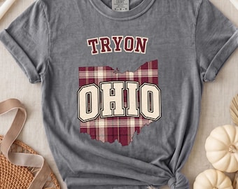 以下が含まれることがあります： チャコールグレーの半袖Tシャツ。マルーンカラーで「TRYON」の文字が入り、その下にチェック柄のオハイオ州の輪郭が描かれています。チェック柄のグラフィックの中央には、クリーム色の大きなブロック体で「OHIO」と記されています。