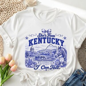 A white t-shirt with a graphic design depicting a horse, flowers, and a state map, along with the text "She's from Kentucky" and "I can tell." The t-shirt is placed on a white surface, with a beige woven basket and some pink flowers nearby.