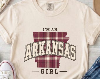 以下が含まれることがあります： クリーム色の半袖Tシャツで、アーカンソー州の形をしたマルーンと白のチェック柄のグラフィックが特徴です。胸元には、州の形の上に濃い茶色のカレッジフォントで「I'M AN ARKANSAS GIRL」という文字がプリントされています。