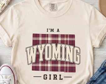 May include: A cream-colored short-sleeve t-shirt featuring a maroon and white plaid square graphic. The text reads I'M A WYOMING GIRL in bold, dark brown block lettering. The shirt has a crew neckline and a Comfort Colors brand label.