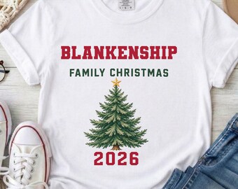 以下が含まれることがあります： 赤い文字でBLANKENSHIP、緑の文字でFAMILY CHRISTMAS、頂上に金の星がある緑のクリスマスツリーのイラスト、そしてツリーの下に赤い文字で2026とプリントされた、前面にデザインがある白い半袖Tシャツ。