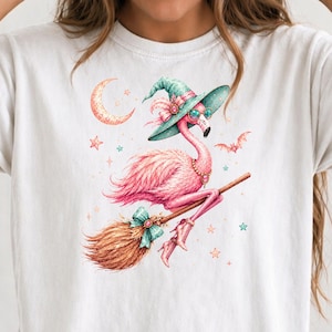 May include: A white t-shirt featuring a graphic of a pink flamingo wearing a teal witch hat, round glasses, a beaded necklace, and lace-up boots. The flamingo is riding a brown broomstick, surrounded by a crescent moon, stars, and a bat.