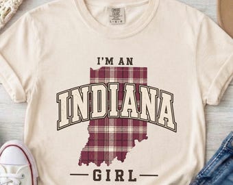 May include: A cream-colored short-sleeve t-shirt featuring a maroon and white plaid graphic of the state of Indiana. The text reads I'M AN INDIANA GIRL in a dark brown collegiate font. The shirt has a crew neckline and a Comfort Colors brand label.