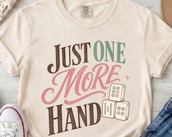 以下が含まれることがあります： クリーム色の半袖Tシャツで、茶色と落ち着いた緑色の文字で「JUST ONE MORE HAND」と書かれています。テキストの下には、3つの麻雀牌がピラミッド状に積み重なったグラフィックイラストが描かれています。
