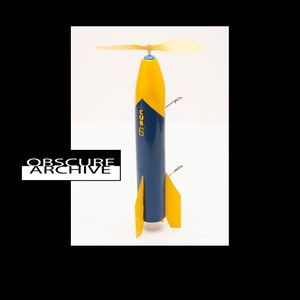 May include: A vintage wooden toy rocket with a yellow and blue color scheme. The rocket has a propeller on top and fins at the base. The text "CUB 6" is printed on the side. The rocket is a nostalgic item.