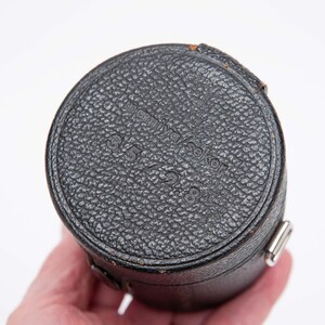 Puede incluir: Un estuche negro y cil&iacute;ndrico para lentes de c&aacute;mara con una superficie texturizada. La parte superior del estuche tiene el texto "minolta seko" y "3.5 / 2.8" en relieve. El estuche tiene un cierre de metal plateado.