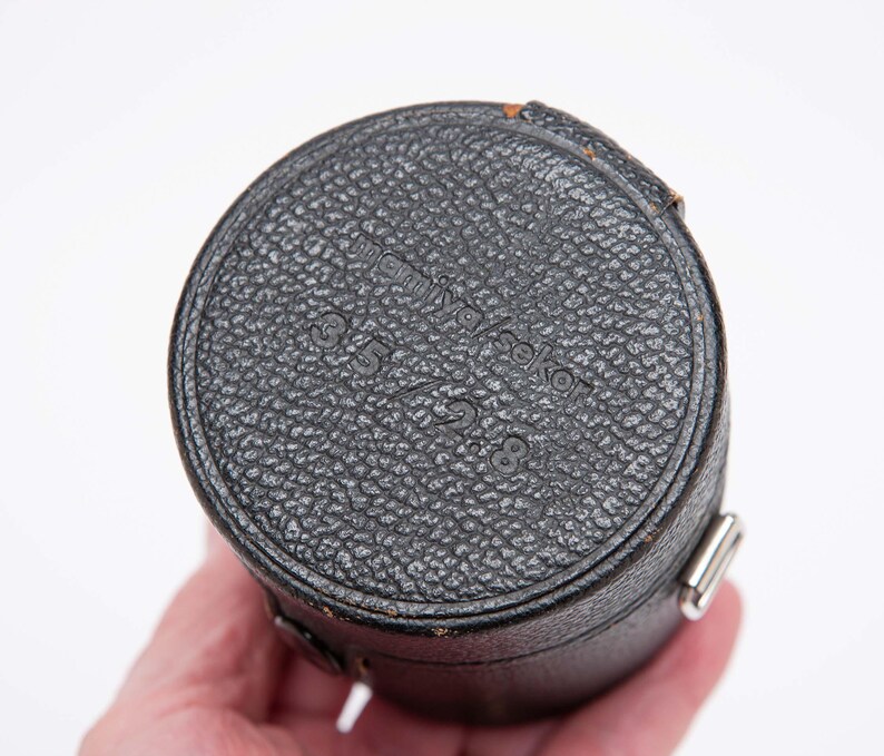 Puede incluir: Un estuche negro y cil&iacute;ndrico para lentes de c&aacute;mara con una superficie texturizada. La parte superior del estuche tiene el texto "minolta seko" y "3.5 / 2.8" en relieve. El estuche tiene un cierre de metal plateado.
