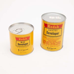 May include: Two yellow Kodak developer cans with red and black text. One can is labeled "D-11 Developer" and the other "Dektol Developer". Both cans are 1 U.S. gallon size and have silver lids.