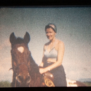 May include: Vintage photograph of a woman riding a brown horse. The woman is wearing a light blue top and a dark skirt. The background shows a field and a cloudy sky. The image has a grainy texture.