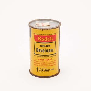 May include: Vintage Kodak DK-50 Developer can. The yellow can features red and black text, including "Kodak" and "Developer." It holds 1 U.S. gallon (3.8 liters). The can has a silver lid and a small paper tag.