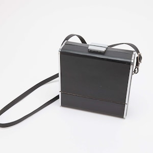 May include: Black rectangular handbag with a silver-tone clasp and trim. The bag has a long, thin black strap for carrying over the shoulder. The bag is made of a shiny material.