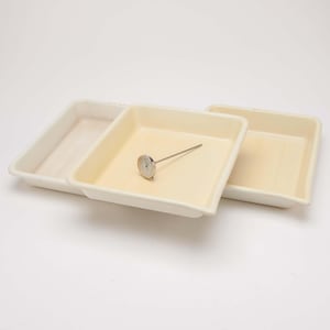 May include: Three off-white, square ceramic trays are stacked on a white surface. A small, silver thermometer is placed inside the middle tray. The trays have slightly rounded edges and a smooth, matte finish. The background is plain white.