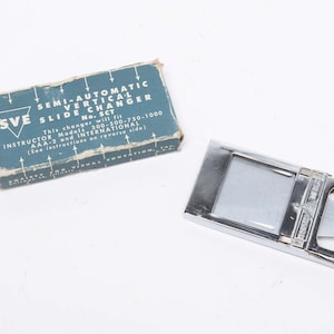 May include: A vintage semi-automatic vertical slide changer with a silver metal frame and a blue box. The box has text that reads "SEMI-AUTOMATIC VERTICAL SLIDE CHANGER No. SCT". The changer is designed for instructor models.