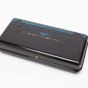 May include: A rectangular black box with a hinged lid. The box has a shiny finish and is decorated with blue and gold accents. The text "THE HULLEBERE RYLE MOPTOPIES" is printed on the lid.