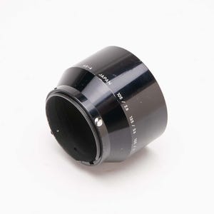 May include: Black lens hood with the text "HS-4 JAPAN 105 / 2.5 135 / 3.5". The lens hood is cylindrical with a flared end. The lens hood is made of a glossy black material.