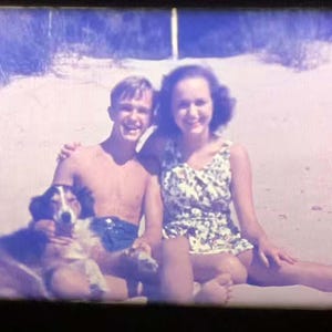 May include: Vintage photograph of a smiling couple and a dog on a sandy beach. The woman wears a floral print swimsuit. The man is shirtless. The dog is brown, white, and black.
